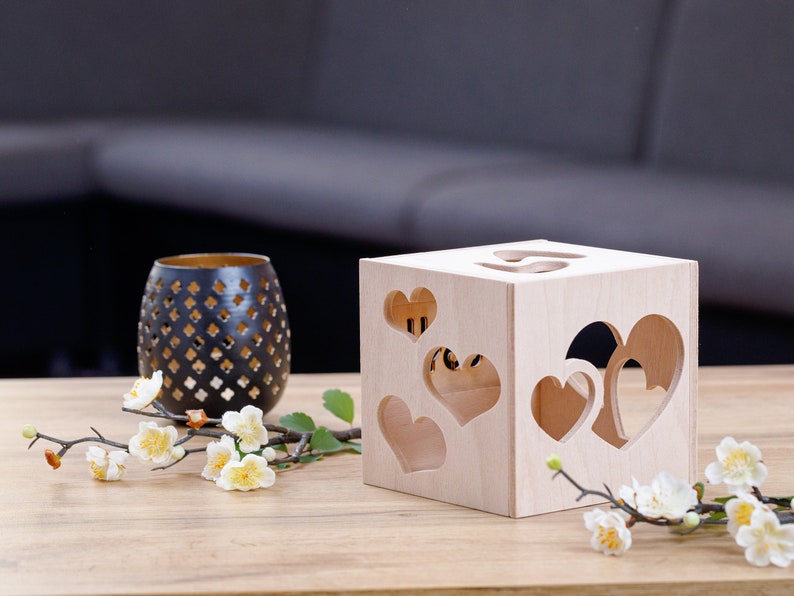 May include: A wooden box with heart-shaped cutouts. The box is light brown and has a natural wood finish. The box is sitting on a wooden table with a branch of white flowers.