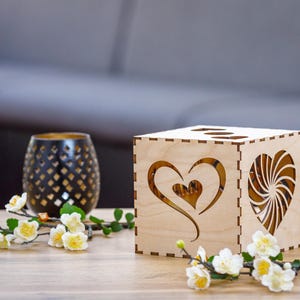 Op de afbeelding: Een houten doos met een hartvormige uitsparing aan de ene kant en een swirl-ontwerp aan de andere kant. De doos is lichtbruin en heeft een natuurlijke houten afwerking. Het staat op een houten tafel met een klein boeket witte bloemen.