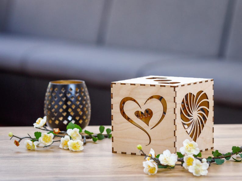 Op de afbeelding: Een houten doos met een hartvormige uitsparing aan de ene kant en een swirl-ontwerp aan de andere kant. De doos is lichtbruin en heeft een natuurlijke houten afwerking. Het staat op een houten tafel met een klein boeket witte bloemen.