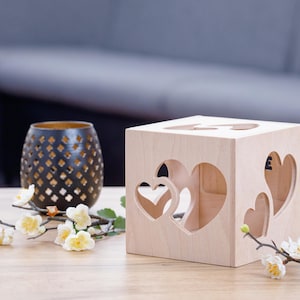 May include: A light wood cube with heart-shaped cutouts. The cube is sitting on a wooden table with a small branch of white flowers and a black metal candle holder.