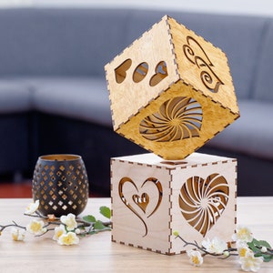 May include: Two wooden cube-shaped candle holders with heart cutouts. One cube is balanced on top of the other. A black decorative candle holder and floral arrangement are on the table. The cubes are made of light-colored wood.