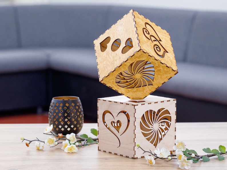 May include: Two wooden cube-shaped candle holders with heart cutouts. One cube is balanced on top of the other. A black decorative candle holder and floral arrangement are on the table. The cubes are made of light-colored wood.