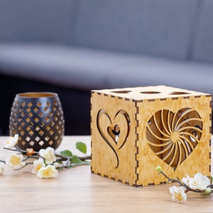 May include: A wooden candle holder with heart-shaped cutouts and a decorative black vase. The candle holder is a cube shape with four holes on top. White flowers and greenery are arranged on the table.