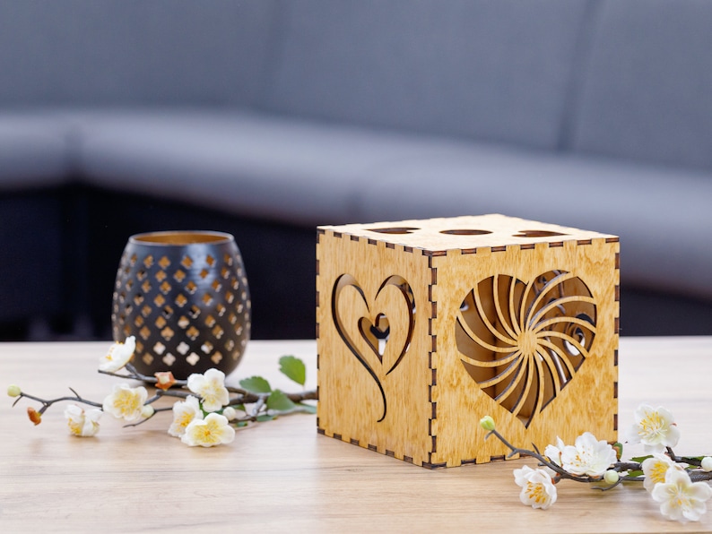May include: A wooden candle holder with heart-shaped cutouts and a decorative black vase. The candle holder is a cube shape with four holes on top. White flowers and greenery are arranged on the table.