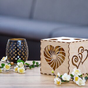 May include: A wooden candle holder with heart-shaped cutouts and a decorative black glass candle holder. The candle holder is made of light-colored wood and has a geometric design. White flowers and green leaves are in the foreground.