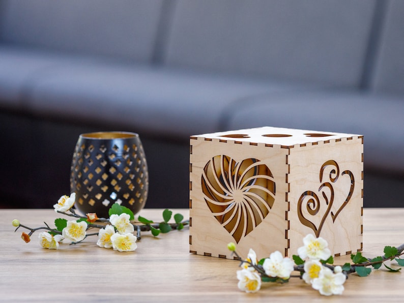 May include: A wooden candle holder with heart-shaped cutouts and a decorative black glass candle holder. The candle holder is made of light-colored wood and has a geometric design. White flowers and green leaves are in the foreground.