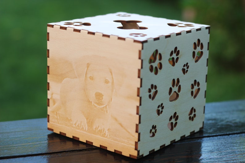 May include: A wooden box with a laser-engraved image of a dog and paw prints on the sides. The box is light brown and has a cutout in the top.