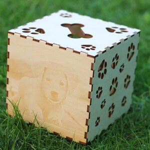 May include: A wooden box with a dog paw print design. The box has a cutout of a bone on the top and a photo of a dog on the front. The box is sitting on a bed of green grass.