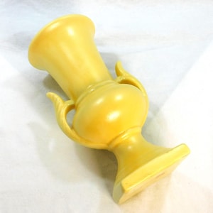 May include: A yellow ceramic vase with two handles. The vase has a rounded body and a square base.