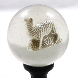 May include: A clear glass ball with a silver alpaca figurine inside. The ball is on a black stand.