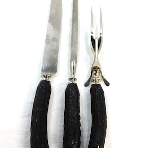 May include: A set of three silver-toned carving utensils with black, textured handles. The set includes a carving knife, a carving fork, and a carving tool.