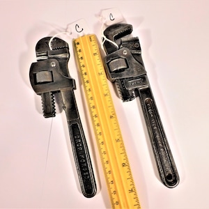 May include: Two vintage adjustable wrenches, one with a black handle and the words "DROP FORGED" on it, and the other with a silver handle and the word "TRUCO" on it. Both wrenches are next to a wooden ruler.