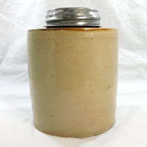 May include: A cylindrical stoneware jar with a metal lid. The jar is a light tan color with a slightly rough texture. The metal lid is silver and sits on top of the jar. The jar is likely used for storage.