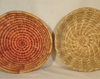 May include: Two woven baskets, one with a natural straw color and the other with a reddish-brown color. Both baskets have a circular shape and a spiral pattern.