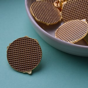 Könnte beinhalten: Nahaufnahme einer goldfarbenen Metallnadel mit einem Waffelmuster. Die Nadel liegt auf einer hellblauen Oberfläche.