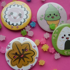 Op de afbeelding: Vier ronde pinnen met cartoon gezichten en Japanse voedselontwerpen. De pinnen bevinden zich op een roze achtergrond met kleine, kleurrijke snoepjes. De ontwerpen omvatten een groene ijshoorn, een bruin blad, een roze swirl en een witte rijstbal.