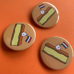 Può includere: Tre bottoni rotondi marroni con un'illustrazione di cartone animato di un dolce sormontato da una bandiera olandese. Il dolce è giallo con una crosta marrone e un cucchiaio di panna montata bianca.