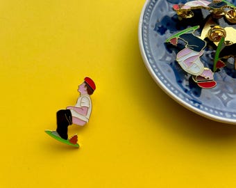 May include: A set of enamel pins featuring a person in a white shirt and red hat sitting on a skateboard. The pins are on a blue and white patterned plate.