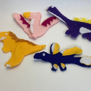 May include: Four felt dinosaur toys. One is pink with purple wings, one is yellow, one is purple with yellow spikes, and one is blue with yellow wings.