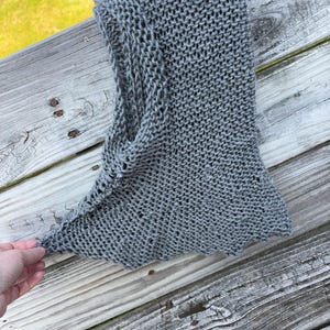 May include: A grey knitted hood made with a loose, open-stitch pattern. The garment features a pointed top and a wide opening for the face, constructed from a single piece of yarn with a textured, hand-knit appearance and a soft, flexible drape.