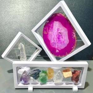 May include: A collection of colorful crystals and gemstones displayed in white, square, floating frames. The collection includes a large, pink and white agate slice, clear quartz crystals, and a variety of smaller stones in shades of purple, blue, green, yellow, and brown.