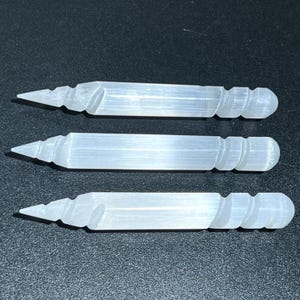 May include: Three clear selenite wands are displayed against a dark background. Each wand has a pointed tip on one end and a rounded, textured end on the other. The wands are approximately 6 inches long.