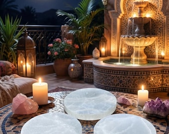 May include: A nighttime scene with a mosaic table featuring three round, white selenite charging plates. Pink and purple crystals, and lit candles surround the plates. A fountain and full moon are in the background, creating a serene ambiance.