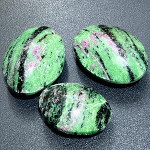 May include: Three oval-shaped zoisite stones. The stones have a green base color with black and pink veining. The stones are polished and smooth, with a natural, organic appearance.