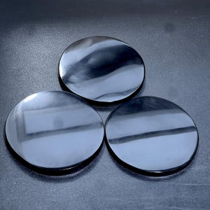 Puede incluir: Tres piedras negras, circulares y pulidas están dispuestas sobre una superficie oscura. Las piedras tienen un acabado reflectante y brillante, y sus superficies lisas captan la luz, creando reflejos y sombras. Las piedras están espaciadas uniformemente.