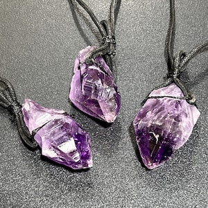 May include: Three amethyst crystal pendants on black cords. The crystals are pointed and have a deep purple color.