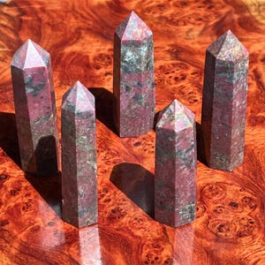 Op de afbeelding: Vijf gepolijste stenen torens met zeshoekige bases en puntige toppen. Elke toren heeft een gevlekt patroon van donkerroze, donkergroene en grijze minerale insluitingen over het hele oppervlak, wat zorgt voor een unieke natuurlijke textuur per stuk.
