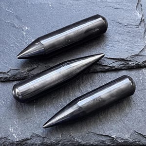 May include: Three polished black stone wands with a smooth, cylindrical body and a tapered, pointed end. Each piece features a dark, reflective surface with subtle grey striations, showcasing a uniform shape and a glossy, finished texture.
