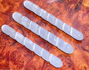 May include: Three translucent white selenite wands, each featuring a distinct spiral or twisted carved pattern along its length. These cylindrical crystal wands have rounded ends and a smooth, polished surface that reflects light across their structure.