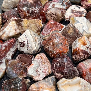 May include: A collection of tumbled agate stones in various shades of red, brown, white, and gray. The stones have a banded pattern and a smooth, polished finish.