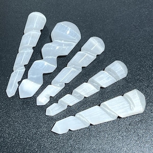 May include: Five white selenite wands with a spiral design. Each wand has a rounded top and a pointed bottom. The wands are arranged on a dark surface, showcasing their translucent quality and unique shape.