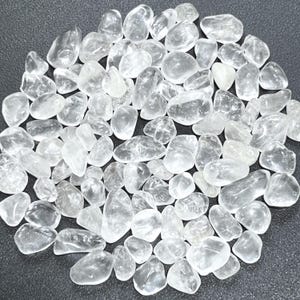 May include: A collection of clear quartz tumbled stones. The stones are smooth, rounded, and vary in size. They are translucent and appear to be polished. The stones are scattered on a dark surface.