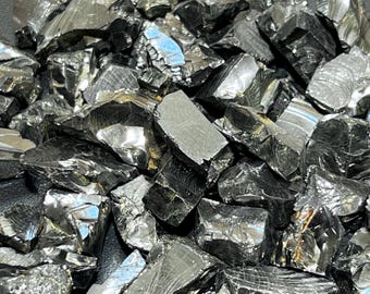 Könnte beinhalten: Eine Ansammlung von rohen, metallisch wirkenden Mineralsplittern mit einer dunklen, reflektierenden und anthrazitfarbenen Oberfläche. Die unregelmäßigen, kantigen Stücke weisen scharfe Kanten sowie glatte, glänzende Facetten auf, die das Licht reflektieren.