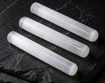 Puede incluir: Tres varitas cilíndricas de selenita con una apariencia fibrosa, blanca y translúcida. Cada varita presenta una superficie lisa y pulida con sutiles estrías verticales a lo largo de la piedra, creando un brillo suave y nacarado.