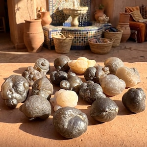 May include: A collection of various-sized, irregularly shaped, polished stones in shades of gray, brown, and cream. The stones are arranged on a terracotta surface, with a blurred background featuring a fountain and decorative elements.