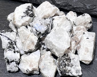 May include: A collection of raw moonstone mineral specimens featuring opaque white and translucent crystalline structures with distinct black tourmaline inclusions. These rough, unpolished stones exhibit natural jagged edges and varied shapes.