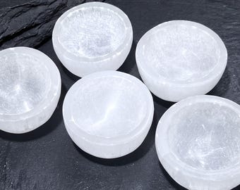 May include: Five small, circular bowls carved from translucent white selenite crystal. Each bowl features a smooth, polished rim and a hollowed-out center, showcasing the natural fibrous texture and crystalline structure of the white stone material.