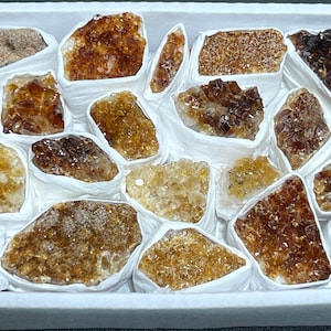 May include: A collection of 18 raw, natural, brown and orange citrine crystals, each wrapped in white tissue paper and arranged in a white box.