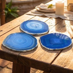 May include: Three blue agate coasters with silver edges are displayed on a wooden table. The coasters have a natural, layered appearance with varying shades of blue. The table is made of wood.