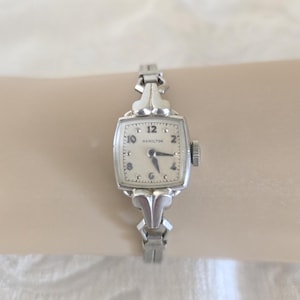 May include: A silver-toned Hamilton wristwatch featuring a square face with a cream-colored dial, black Arabic numerals, and dark hands. The watch has a decorative metal link band with a polished finish and a small winding crown on the side.