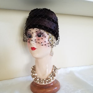 May include: A vintage dark brown hat with a net veil, displayed on a mannequin head. The mannequin wears a pearl and rhinestone necklace. The hat has a rounded crown and a fine net veil that drapes over the face.