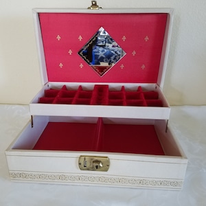 May include: A vintage white jewelry box with a red interior. The box has two levels, a mirror, and gold-colored hardware. The interior is lined with red velvet and has compartments for storing jewelry.
