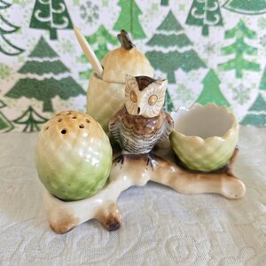 May include: A ceramic owl-themed salt and pepper set with a matching sugar bowl and egg cup. The owl is perched on a branch, and the set has a green and brown color scheme. The background features a Christmas tree pattern.
