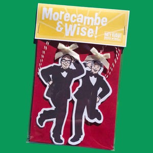 May include: Two die-cut paper ornaments featuring illustrations of Morecambe and Wise in black tuxedos, each topped with a small cream ribbon bow and red and white twine, packaged on a red card with a yellow header reading Morecambe & Wise!