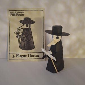 Puede incluir: Una figura de Doctor de la Peste de papel con sombrero, capa y pico negros, sosteniendo un lazo blanco y un bastón de madera. Junto a ella, una tarjeta con el texto "Hey Kids Rock'n'Roll Folk Fancies" y "5. Plague Doctor".