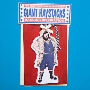 Puede incluir: Decoración colgante roja y blanca con una ilustración de un luchador. El texto dice: "¡PARA LOS FANS DE LA LUCHA LIBRE EN TODAS PARTES! DECORACIÓN COLGANTE GIANT HAYSTACKS". Se adjuntan un lazo blanco y una cuerda azul y blanca.